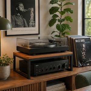 May include: A black turntable with a clear dust cover sits on a wooden and black metal stand. Beneath the stand is a black Denon audio amplifier. To the right, a Miles Davis Kind of Blue vinyl record is displayed in a rack.