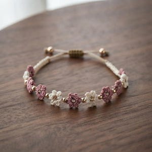 Puede incluir: Una pulsera con motivos de flores de ganchillo en colores malva y crema alternados, unidos por pequeñas cuentas metálicas doradas. La banda está hecha de un cordón de color claro con un mecanismo de cierre de nudo corredizo en la parte posterior.