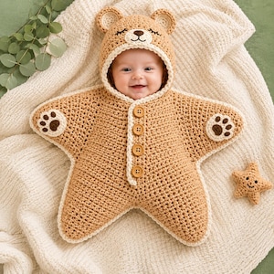 Peut inclure: Une gigoteuse en crochet en forme d'étoile de couleur beige avec une capuche ornée d'oreilles et d'un visage d'ours. Le vêtement présente quatre boutons en bois sur le devant, une bordure crème et des pattes d'ours marron foncé.