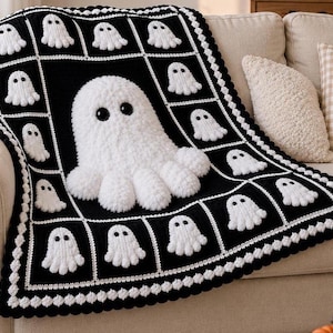 May include: A black and white crochet blanket featuring a grid pattern of small white ghost motifs. A large, three-dimensional white ghost with black eyes is centered on the blanket. The edges are finished with a scalloped white crochet border.