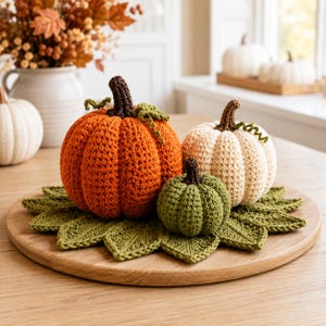 Può includere: Tre zucche all'uncinetto nei colori arancione, crema e verde, ciascuna con un gambo marrone scuro e piccoli viticci ricci. Le zucche sono disposte su un tappetino all'uncinetto verde a forma di foglia, posizionato su un tagliere di legno rotondo.
