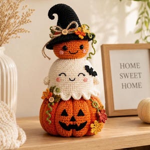 Peut inclure: Une décoration d'Halloween en crochet empilé composée d'une grande citrouille orange, d'un fantôme blanc au milieu et d'une petite citrouille orange portant un chapeau de sorcière noir. L'ensemble est orné de petits boutons en bois et de feuilles.