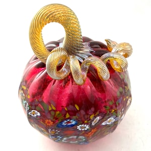 May include: A hand-blown glass pumpkin in a deep red hue, featuring a ribbed surface decorated with colorful floral millefiori patterns. The top is adorned with a clear, spiraled, amber-toned glass stem that curls into a decorative vine.