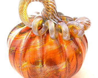 May include: A decorative glass pumpkin with an orange body and a swirling, transparent stem. The pumpkin has a textured surface with streaks of gold and other colors. The stem is a light amber color and is curled.