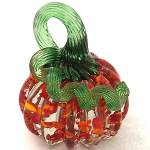 May include: A decorative glass pumpkin sculpture featuring a clear body with embedded red, orange, and yellow flecks. The top includes a prominent, spiraled green glass stem and a curled green glass vine extending across the ribbed surface.