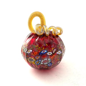 May include: A decorative glass pumpkin with a red base and colorful floral patterns. The pumpkin has a gold-colored, swirled stem. The glass pumpkin is a unique fall or autumn home decor item.