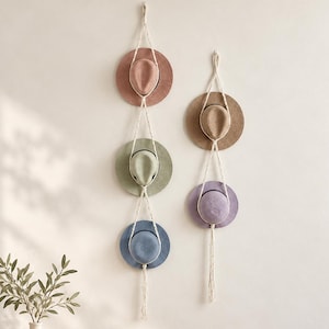 May include: Two vertical macrame wall hangers made of off-white cord, each holding multiple hats. The left hanger holds three hats in dusty pink, sage green, and blue. The right hanger holds two hats in tan and lavender.