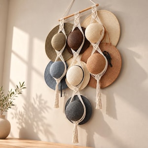 May include: A macrame wall hanging made of off-white cord suspended from a wooden dowel, featuring three vertical strands that hold seven circular hats in various shades of brown, tan, blue, and black, with tasseled ends.