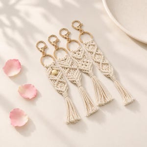 May include: Four off-white macrame keychains featuring intricate knotted patterns, gold-toned metal lobster clasps, and dangling tassels. One keychain includes a single round, light-coloured bead integrated into the woven cord design.