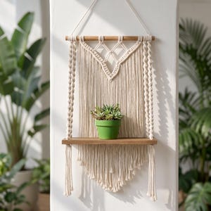 May include: A wall-hanging macrame shelf featuring off-white cotton cord knotted in intricate patterns, suspended from a horizontal wooden dowel. A flat wooden board serves as a shelf, holding a small green pot containing a cluster of succulents.