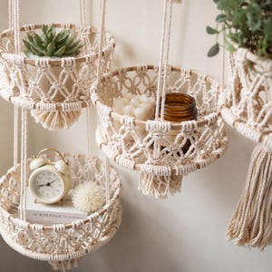 May include: A set of hanging macrame baskets made from off-white cotton cord with wooden circular frames. The baskets hold various items including a green succulent, a white bubble candle, a brown glass jar, and a white alarm clock on a book.