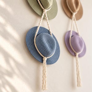 May include: Four straw hats in olive green, tan, blue, and lavender are displayed on a wall. Each hat is suspended by a thick, cream-coloured braided rope that loops around the crown and hangs down in a long, decorative tassel at the bottom.