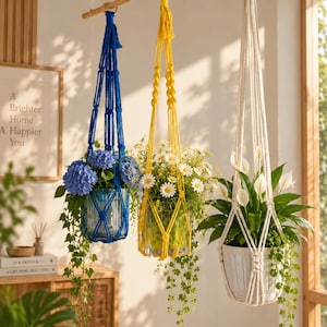 May include: Three macrame plant hangers in blue, yellow, and off-white, suspended from a wooden rod. The blue hanger holds a glass vase with blue hydrangeas, the yellow holds a glass vase with white daisies, and the off-white holds a white pot.