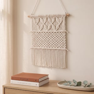 May include: A cream-colored macrame wall hanging featuring a wooden dowel, intricate knotted patterns, and a long fringe. The textile art displays a top section of diamond-shaped knots above a wider band of open lattice weave, finished with hanging cords.