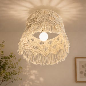 May include: A cream-colored macrame lampshade featuring a woven diamond pattern and long fringe trim along the bottom edge. The shade is suspended from the ceiling and houses a single round white light bulb that casts a soft glow.
