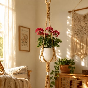 Può includere: Un vaso per piante in ceramica bianca sostenuto da un supporto in macramè color giallo senape con un design annodato e una lunga nappa sul fondo. Il vaso contiene una pianta verde con fiori rossi vivaci, appeso a un anello di legno.