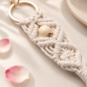 Puede incluir: Un llavero de macramé hecho con cordón de algodón color hueso anudado en un patrón de diamantes con una cuenta de madera lisa y clara centrada en el tejido. El macramé está sujeto a una anilla circular de metal en tono dorado.