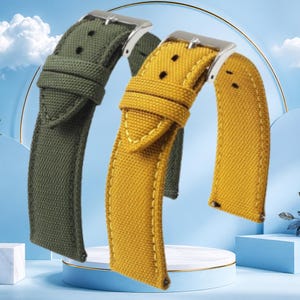 May include: Two watch straps made of textured fabric, one in olive green and one in mustard yellow. Each strap features a silver-toned metal buckle, a matching fabric keeper loop, and visible stitching along the edges for a secure fit.