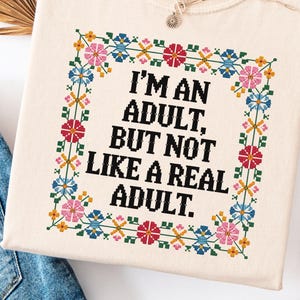 May include: Cream-colored t-shirt with the text "I'M AN ADULT, BUT NOT LIKE A REAL ADULT." The text is framed by a colorful floral cross-stitch design. A silver necklace with a small pendant rests on the shirt.