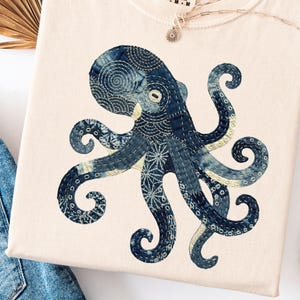 May include: Cream-colored t-shirt featuring a detailed blue octopus design. The octopus is intricately patterned with a mix of textures and shades of blue. The shirt has a crew neck and short sleeves. A silver necklace with a small pendant is on the shirt.