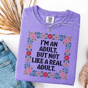 May include: A lavender Comfort Colors sweatshirt with the text "I'M AN ADULT, BUT NOT LIKE A REAL ADULT" in a cross-stitch style design. The design is framed with colorful flowers. A silver necklace with a sun pendant is draped across the shirt.