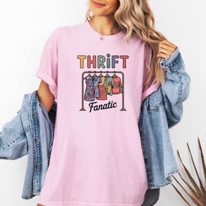 May include: A light pink t-shirt with the words "THRIFT Fanatic" in colorful letters. The graphic features a clothes rack with dresses and tops. The person is wearing a denim jacket.