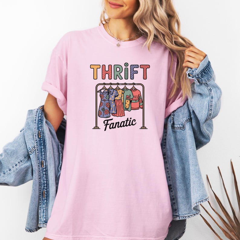 May include: A light pink t-shirt with the words "THRIFT Fanatic" in colorful letters. The graphic features a clothes rack with dresses and tops. The person is wearing a denim jacket.