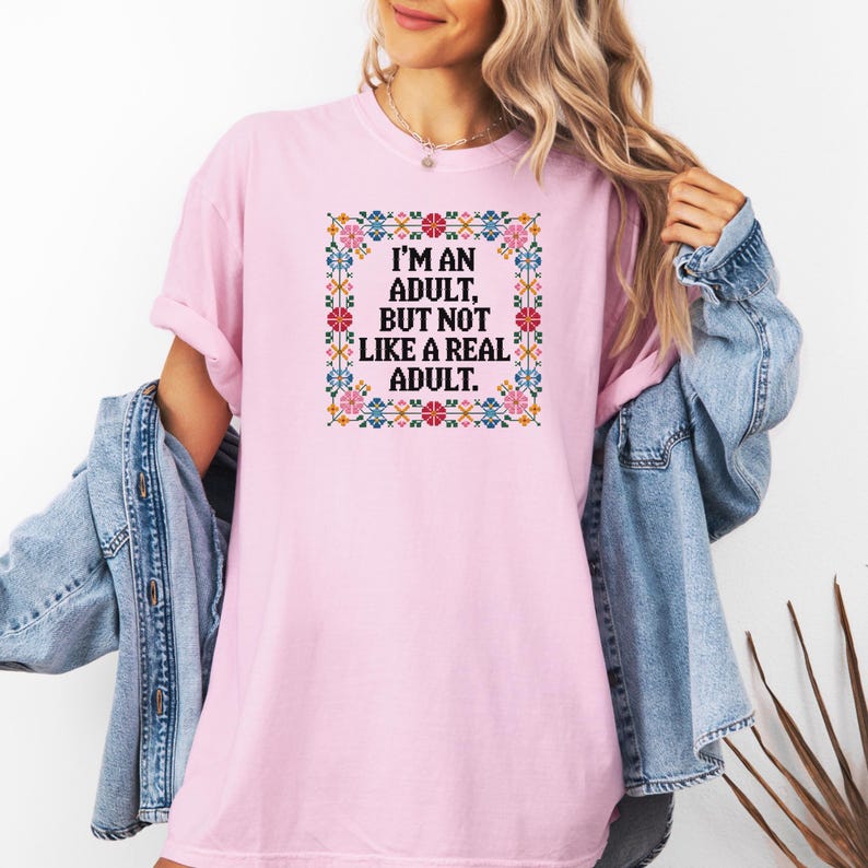 May include: A light pink t-shirt with the text "I'M AN ADULT, BUT NOT LIKE A REAL ADULT" in a square floral design. The design features a variety of colors including pink, blue, yellow, and green. The shirt has short sleeves.