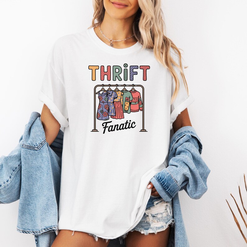 May include: White t-shirt with the word "THRIFT" in colorful letters above a graphic of a clothing rack with dresses and the word "Fanatic" below. The shirt is being worn.