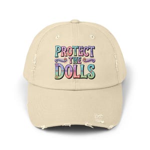 May include: A beige distressed baseball cap with the text "Protect the Dolls" in colorful letters.
