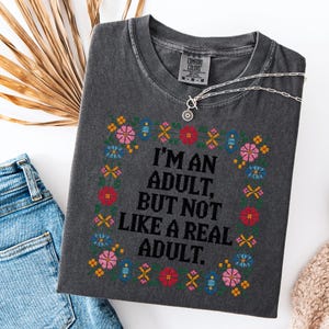 May include: Dark gray Comfort Colors t-shirt with a floral embroidered design and the text "I'M AN ADULT, BUT NOT LIKE A REAL ADULT." A silver necklace and blue jeans are also visible.