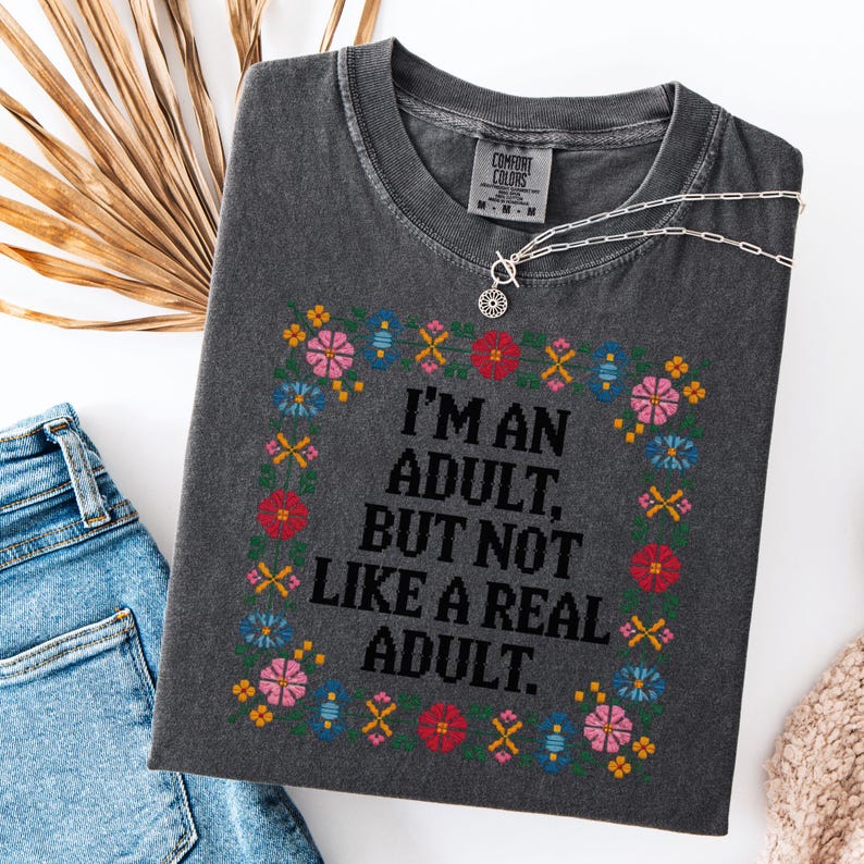 May include: Dark gray Comfort Colors t-shirt with a floral embroidered design and the text "I'M AN ADULT, BUT NOT LIKE A REAL ADULT." A silver necklace and blue jeans are also visible.