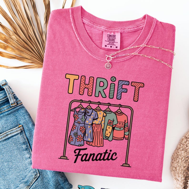 May include: A pink Comfort Colors t-shirt with the word "THRIFT" in orange, yellow, and green letters. Below the word is a graphic of a clothing rack with dresses and tops. The word "Fanatic" is below the clothing rack. A silver necklace is draped across the shirt.
