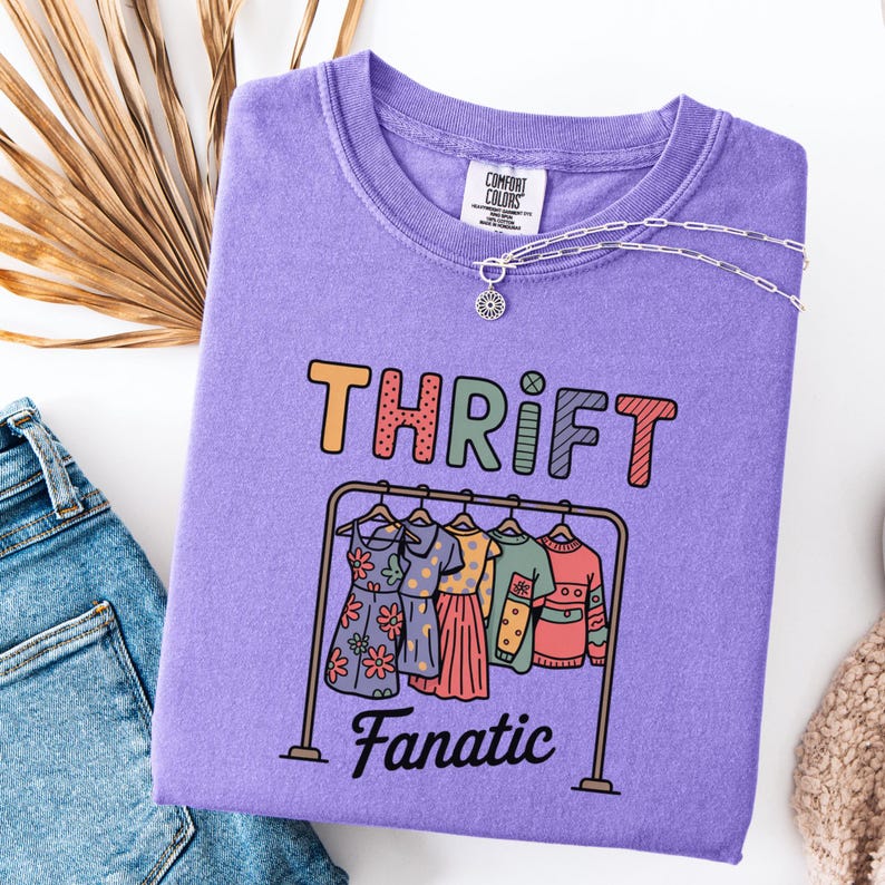 May include: A lilac Comfort Colors sweatshirt with the words "THRIFT Fanatic" in colorful letters. Below the text is a graphic of a clothing rack with dresses and tops. A silver necklace with a sun pendant is on the shirt.