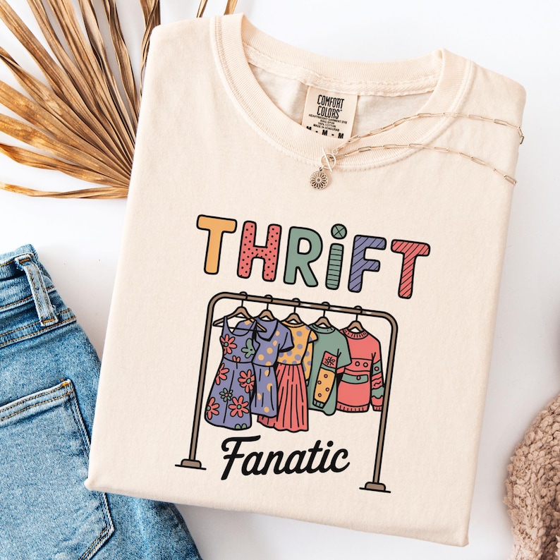 May include: Cream-colored t-shirt with the word "THRIFT" in colorful letters above a cartoon clothes rack with dresses. The word "Fanatic" is below the rack. A gold necklace and a Comfort Colors tag are visible.