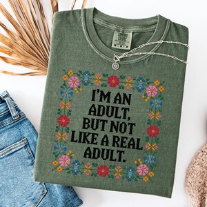 May include: Olive green t-shirt with the text "I'M AN ADULT, BUT NOT LIKE A REAL ADULT" framed by a floral cross-stitch design. A silver necklace rests on the shirt. The shirt is folded, with a pair of blue jeans and a beige sweater in the background.