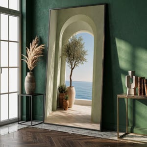 May include: A large, rectangular floor mirror with a thin black frame. The mirror reflects a Mediterranean-style arched balcony with a potted olive tree, a view of the blue ocean, and a clear sky. The mirror stands against a dark green wall.