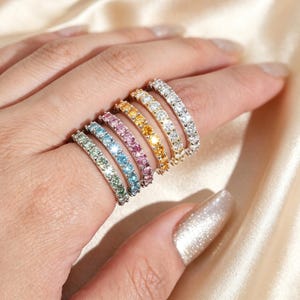 May include: A collection of six thin metal rings, each featuring a single row of small, round, faceted gemstones in different colors including light green, light blue, pink, yellow, orange, and clear, arranged in a parallel stack on a finger.