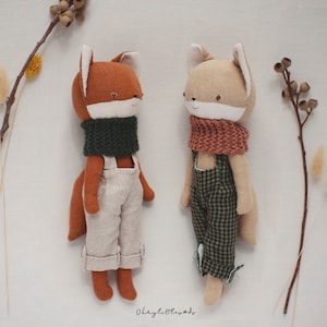 Może przedstawiać: Dwie ręcznie robione pluszowe zabawki w kształcie lisa z haftowanymi twarzami. Jeden lis nosi zielony szalik i beżowe ogrodniczki, drugi nosi różowy szalik i zielono-czarne kratkowane ogrodniczki.