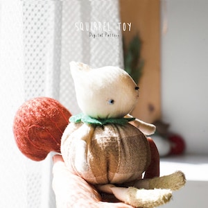 May include: A stuffed squirrel toy with a cream-colored head, a round body featuring a tan and white checkered pattern, a large textured rust-colored tail, and a green felt collar. The text SQUIRREL TOY Digital Pattern is visible above.