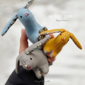 May include: Three handmade Waldorf bunny toys in blue, yellow, and gray fabrics. Each bunny has a floral patterned fabric on the inside of the ears. The toys are held in a hand and the text "WALDORF BUNNY Digital Pattern" is visible above the toys.