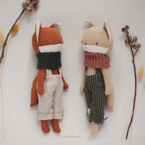 May include: Two plush fox dolls with embroidered facial features. The left fox is rust-colored with a dark green knit scarf and beige overalls. The right fox is tan with a terracotta knit scarf and green checkered overalls. Text reads HushThread.