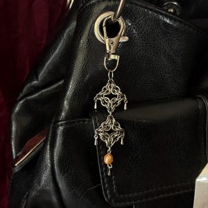 May include: A silver-toned metal chainmaille bag charm featuring two interconnected square-shaped woven links, small dangling metal accents, and a single round peach-coloured bead at the bottom, attached to a silver-toned lobster claw clasp.