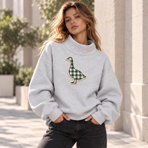 Puede incluir: Una sudadera gris claro de cuello alto con un gran parche central de un ganso con estampado de cuadros vichy verde y blanco. La zona del cuello incluye el nombre EMILY bordado en hilo claro. La prenda tiene mangas largas y puños acanalados.