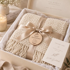 Op de afbeelding: Een crèmekleurige, getextureerde gebreide deken met franjes, verpakt in een champagnekleurig satijnen lint. Een rond houten label bevat de tekst Happy Anniversary A & J 10.14.2023. Een bedankkaartje met de tekst With Gratitude is inbegrepen.