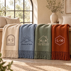 Op de afbeelding: Vier geweven plaids met franjes in crème, blauw, groen en oranje. Elke plaid heeft een uniek gepersonaliseerd ontwerp: een boog met A+J en een datum, een zon boven heuvels met namen, een huisomtrek met een familienaam en een zeshoek met L+M.