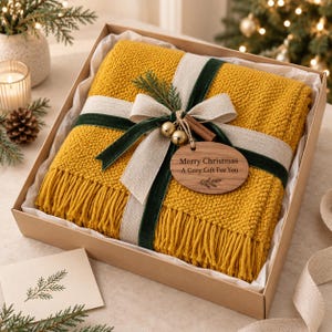 Può includere: Una coperta in maglia color giallo senape con bordi sfrangiati, presentata in una scatola regalo di cartone. La coperta è avvolta da un nastro di lino beige, un nastro di velluto verde scuro, un rametto di pino, due campanellini dorati e un'etichetta in legno.