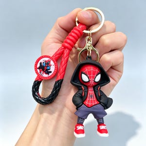 May include: A keychain featuring a stylized character with a red and black spider-web patterned suit, a black hoodie, grey shorts, and red sneakers. The keychain includes a braided red and black wrist strap with a circular charm showing a graphic.
