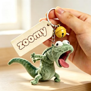 May include: A plush green lizard keyring featuring a wide-open mouth with a pink tongue, large white eyes, and a metal ring attached to a yellow bell and a rectangular wooden tag engraved with the word "ZOOMY" in a bold, rounded font.