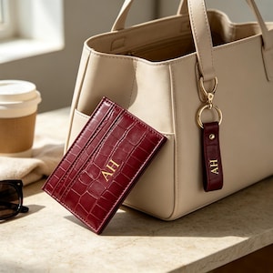 May include: A beige tote bag with a matching beige handle, paired with a dark red crocodile-textured card holder and a matching keychain, both featuring the gold-embossed initials AH. The set is displayed alongside sunglasses and a coffee cup.
